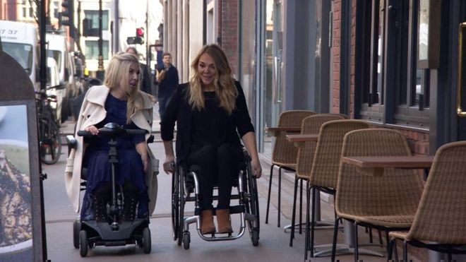 One person using an electric scooter and one person using a wheelchair make their way along a sidewalk, they are conversing. 