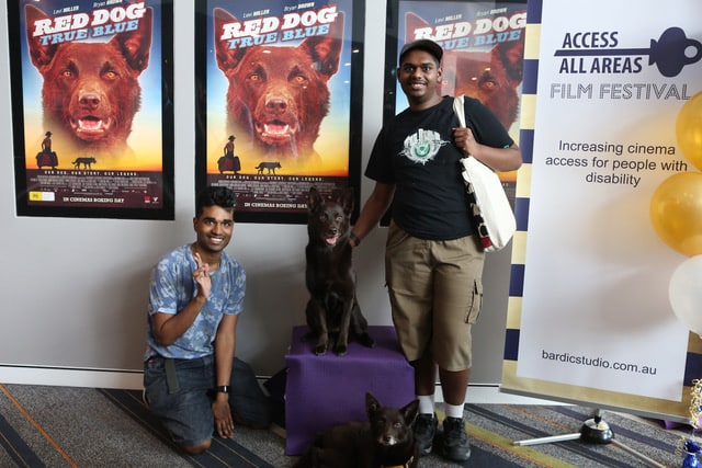 Two people that appear to be from CALD backgrounds stand in front of several Red Dog posters on a wall. One person is patting a dog that is sitting on a podium in front of the posters. There is also a dog at their feet. 