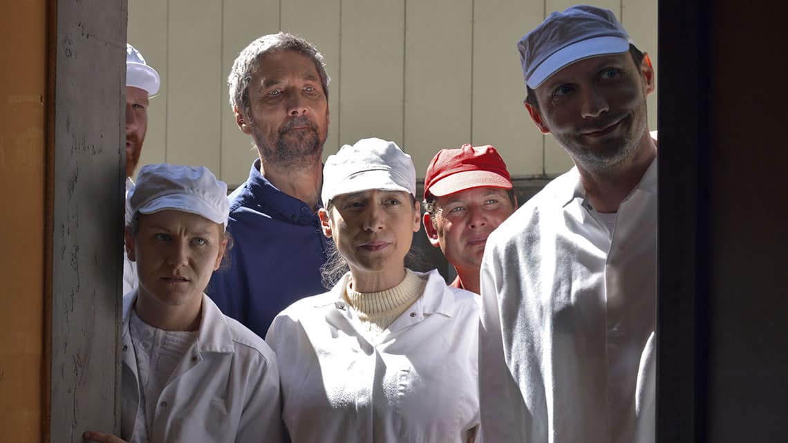 Six people are peering through a doorway. Four of the people are wearing white overalls and white hats. One person is wearing a red hat and one person is wearing a blue shirt and no hat. Each person has a different expression. 