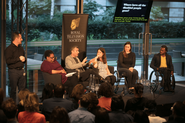 Six people are on a stage addressing an audience. Three people are wheelchair users. One person is standing and is possibly a sign language interpreter.  