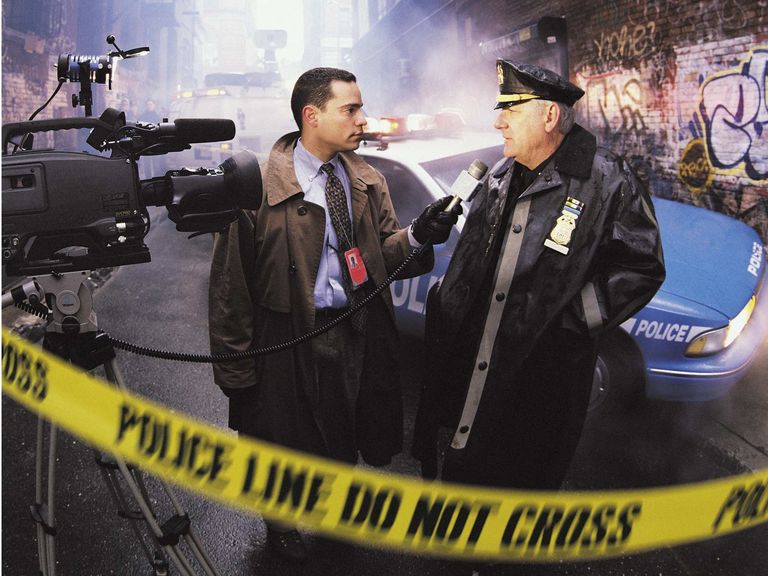 Two people stand in a graffitied alleyway in front of a police car. One person is a police officer, the other is a reporter. The reporter is holding a microphone. There is a camera to the left of screen and caution tape across the entire image saying "Police line do not cross". 