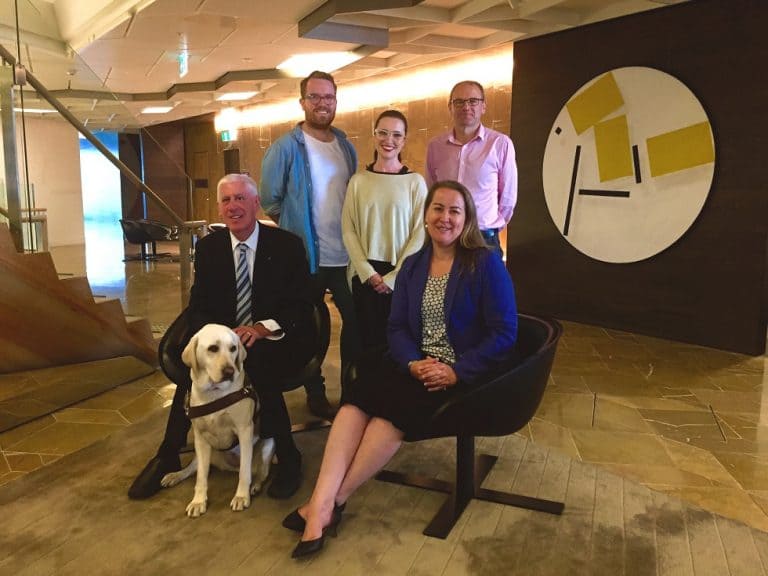 Five people are pictured in a corporate environment. Three people are standing and two are seated. One person is seated next to their guide dog. 