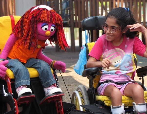 A child and a puppet are seated in individual wheelchairs. The child is smiling.  